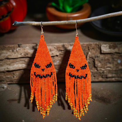 Handmade beaded earrings with an orange jack-o'-lantern face design and long orange fringe, perfect for Halloween-themed outfits. Spooky background 