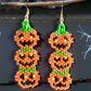 Handmade beaded earrings featuring a vertical stack of three smiling jack-o'-lantern pumpkins in orange, green, and black, perfect for Halloween-themed outfits. Black background 