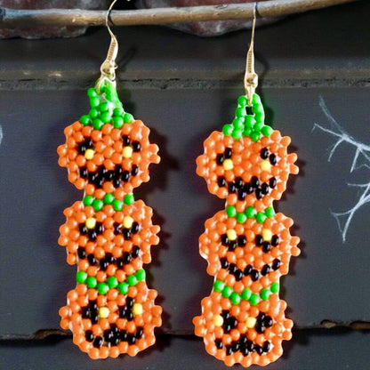 Handmade beaded earrings featuring a vertical stack of three smiling jack-o'-lantern pumpkins in orange, green, and black, perfect for Halloween-themed outfits. Black background 
