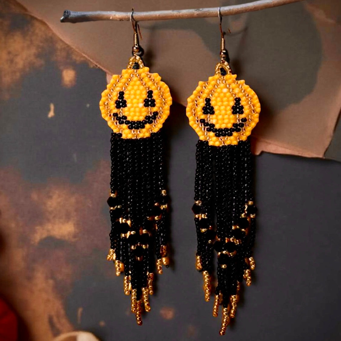 Handmade Halloween beaded earrings with yellow smiley pumpkin faces and long black and gold beaded fringe hanging from hook-style ear wires. Spooky background 