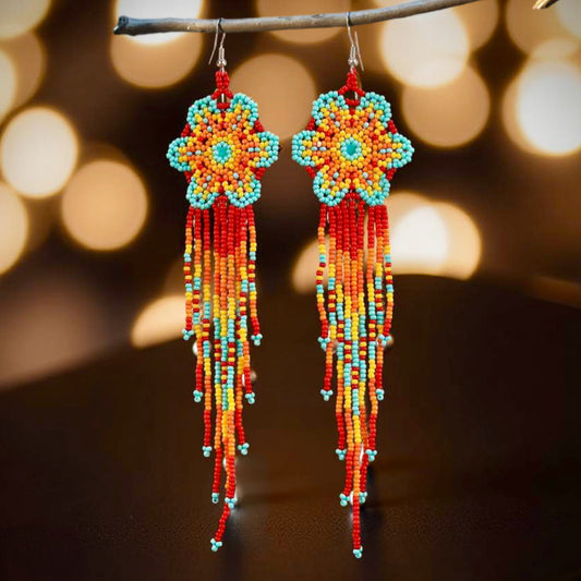 Handmade beaded earrings featuring a flower design with red, orange, yellow, and turquoise beads, complemented by long tassel fringe, with hypoallergenic 316L surgical steel lever backs, set against bokeh lights 