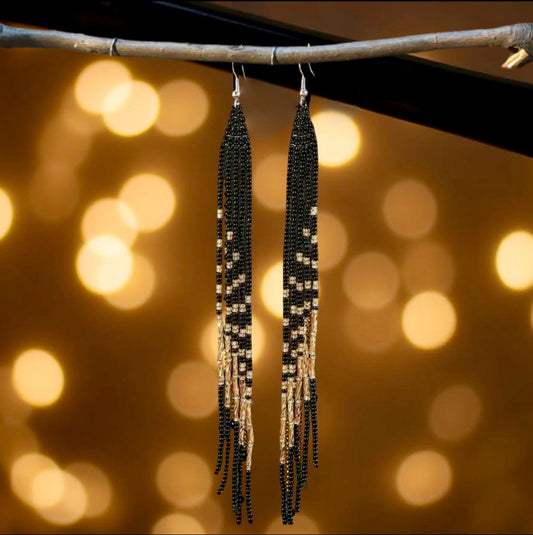 A pair of long handmade beaded earrings with a sleek black and gold fringe design, hanging from hooks against a blurred bokeh golden background.