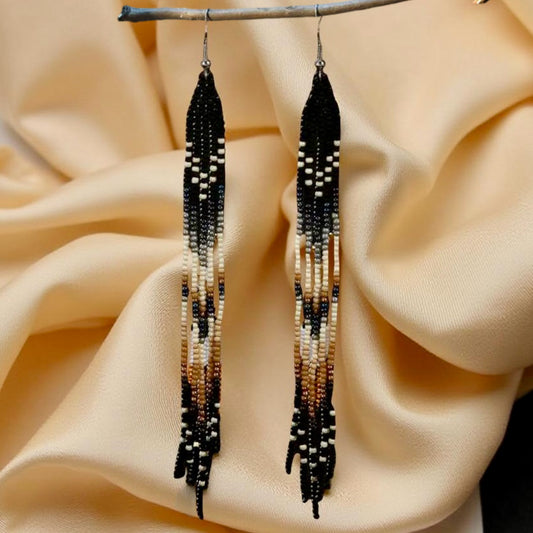 A pair of long handmade beaded earrings featuring a black, cream and tan colour pattern with a flowing fringe design, hanging against a soft beige fabric background.