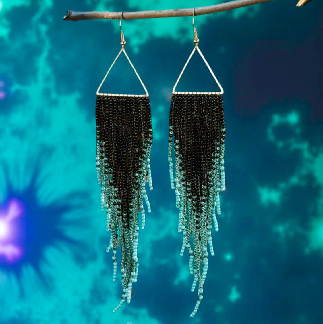 A pair of handmade beaded earrings featuring a triangular top and long black and teal fringe, hanging from hypoallergenic 316L surgical steel lever back hooks in front of blue tie dye background 