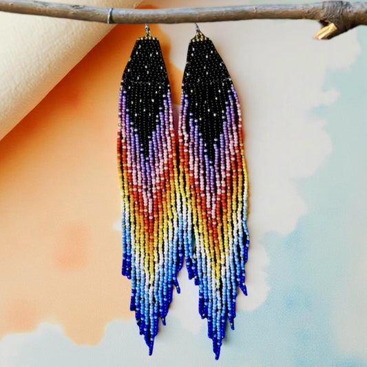 Handmade beaded earrings with a long rainbow fringe, transitioning from black to purple, pink, red, yellow, and blue, hanging from hypoallergenic 316L surgical steel lever backs. Pastel background 