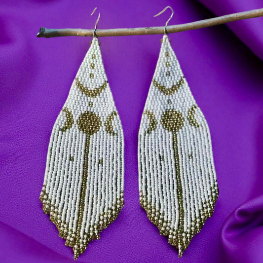 Handmade beaded earrings with a white and gold moon phase design, hanging from a twig against a purple satin background. 