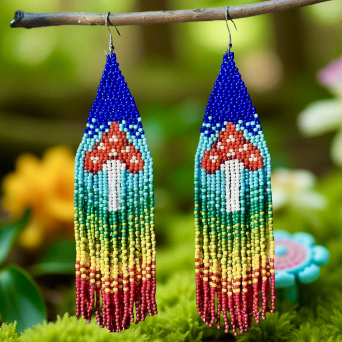 Handmade beaded earrings with a colourful rainbow fringe and red-and-white mushroom design, finished with hypoallergenic surgical steel hooks I front of grass and colourful flowers