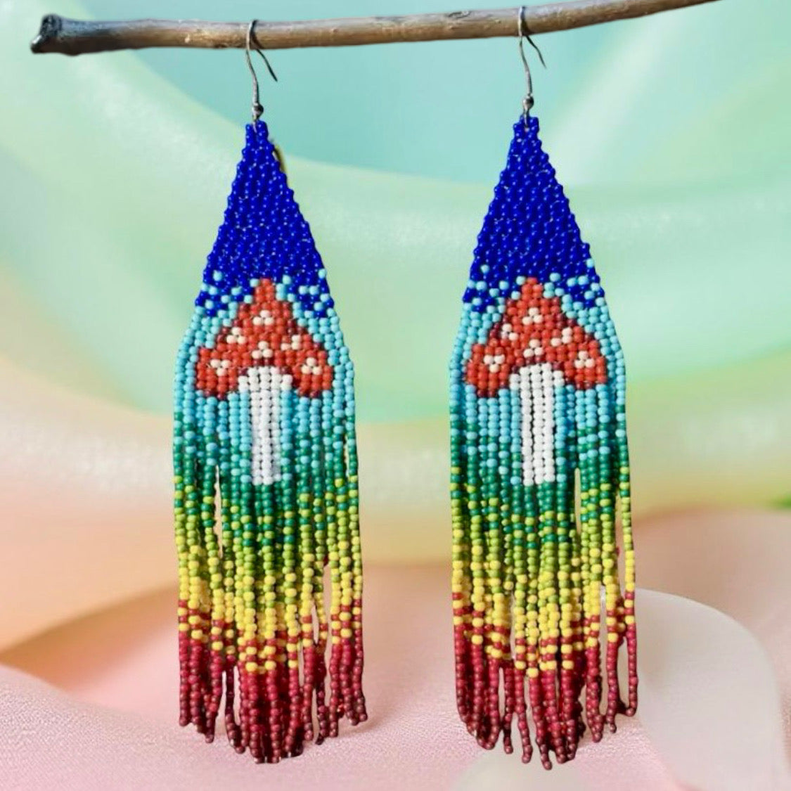 Handmade beaded earrings with a colorful rainbow fringe and red-and-white mushroom design, finished with hypoallergenic surgical steel hooks. In front of a pastel background 