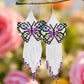 A pair of handmade beaded earrings featuring a multicoloured butterfly design with black accents and a long white fringe, displayed on a floral background.