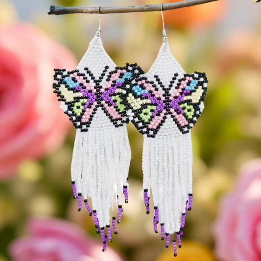 A pair of handmade beaded earrings featuring a multicoloured butterfly design with black accents and a long white fringe, displayed on a floral background.