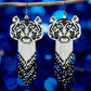 Handmade beaded earrings featuring a white tiger face with blue eyes and a long black, white, and grey beaded fringe, hanging from surgical steel lever backs.