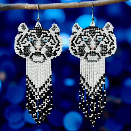 Handmade beaded earrings featuring a white tiger face with blue eyes and a long black, white, and grey beaded fringe, hanging from surgical steel lever backs.