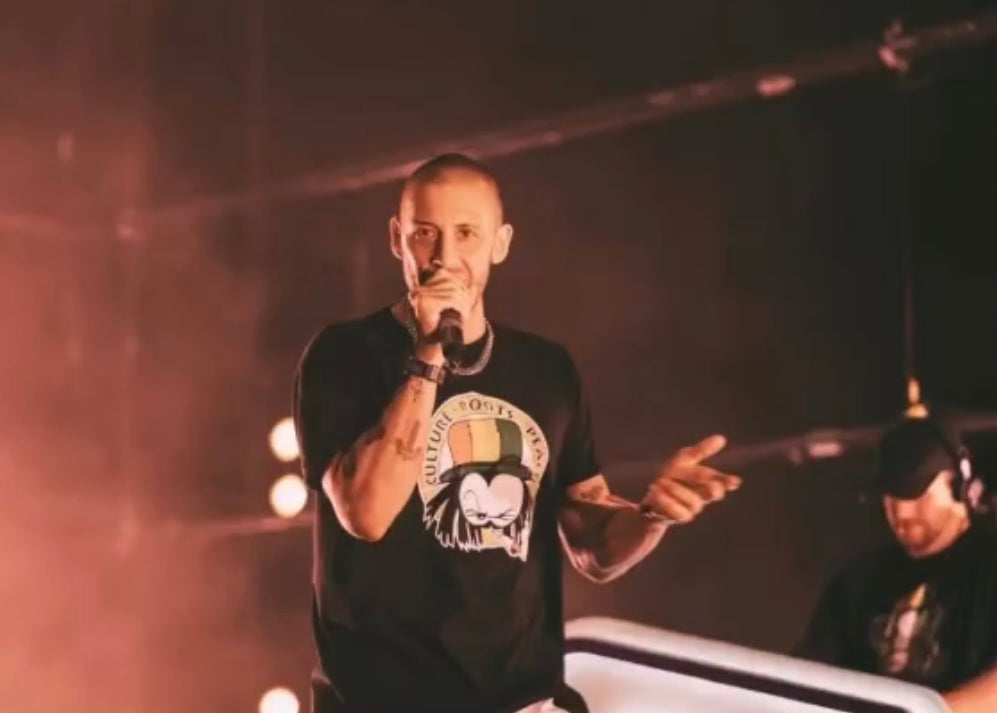 Performer wearing a black Dready Culture Roots Peace graphic T-shirt on stage under warm concert lighting.