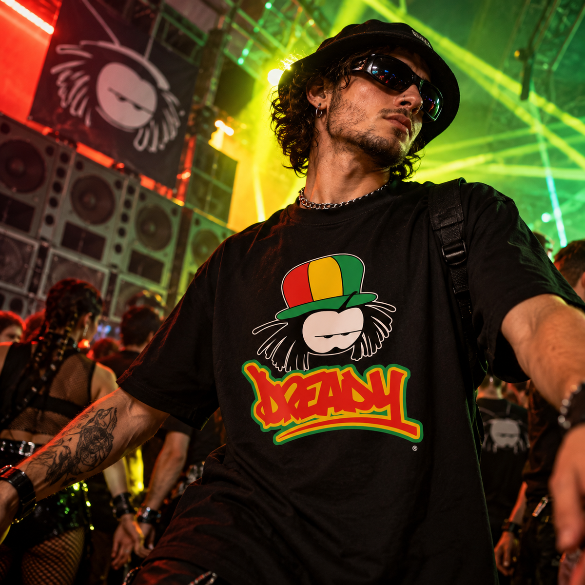 Dreamy black graphic T-shirt featuring a red, yellow and green character design, styled at an underground drum and bass rave with towering speakers and vibrant warehouse lighting.