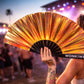 Person holding iridescent festival fan at sunset rave with glowing orange lighting