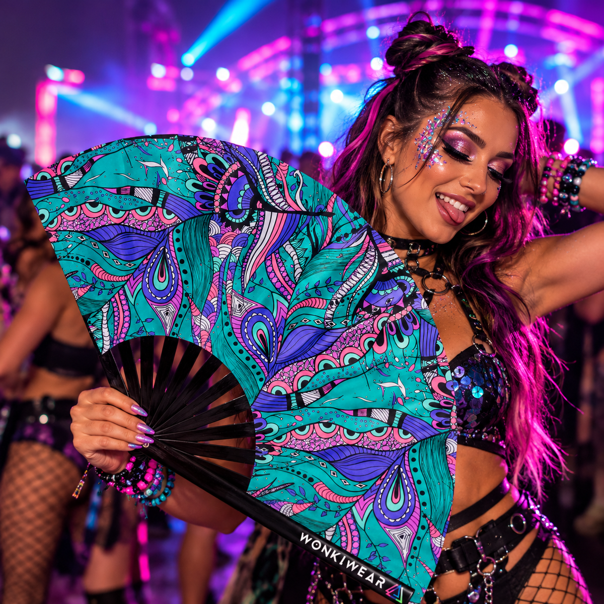 Woman holding the Mystic Paisley XL rave fan by WonkiWear at a neon-lit festival, showcasing its vibrant teal, purple and pink paisley design against colourful rave lights.