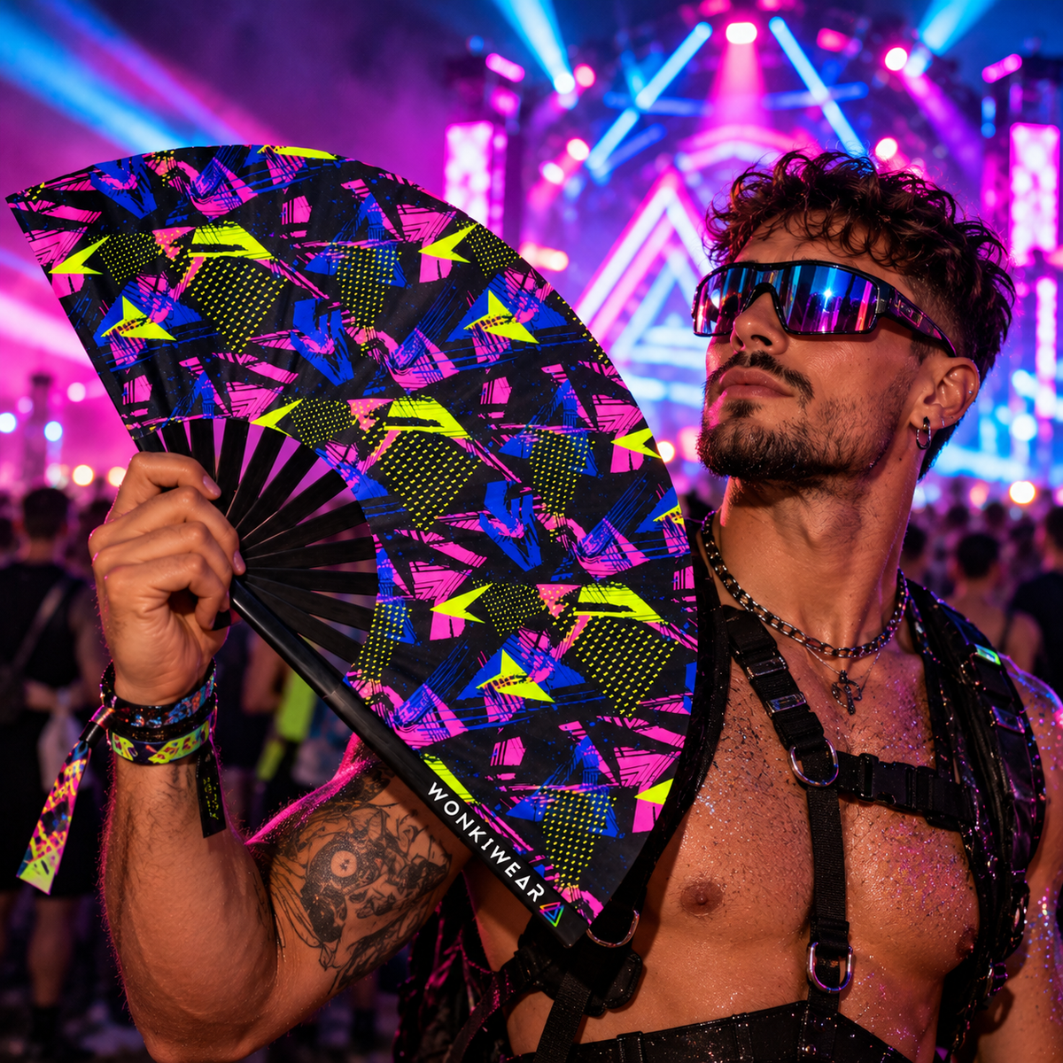 WonkiWear Retro Riot XL folding festival fan featuring bold neon pink, electric blue and acid yellow geometric patterns, held by a male festival-goer wearing mirrored sunglasses and black ravewear at a neon-lit nighttime rave.