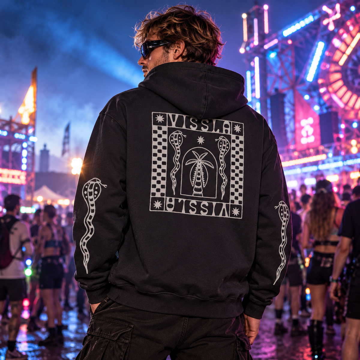 Male raver wearing the black Vissla Offshore Eco PO Hoodie with checkerboard, palm tree and snake graphics at a colourful night festival.
