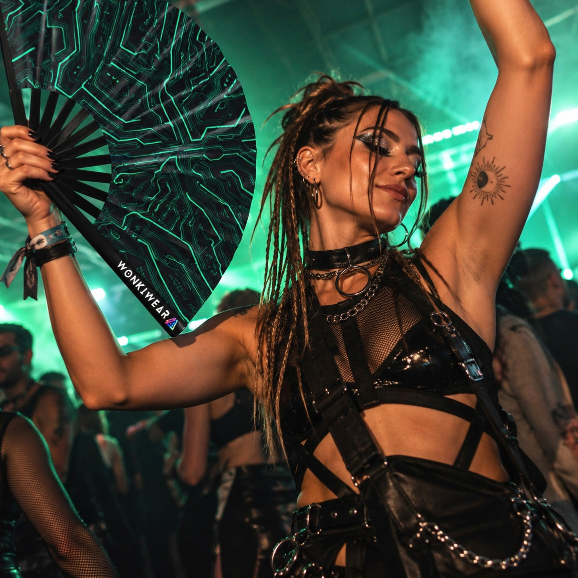 Woman dancing at an underground techno rave holding a black folding fan with teal Cyber Circuit graphics and WonkiWear branding.