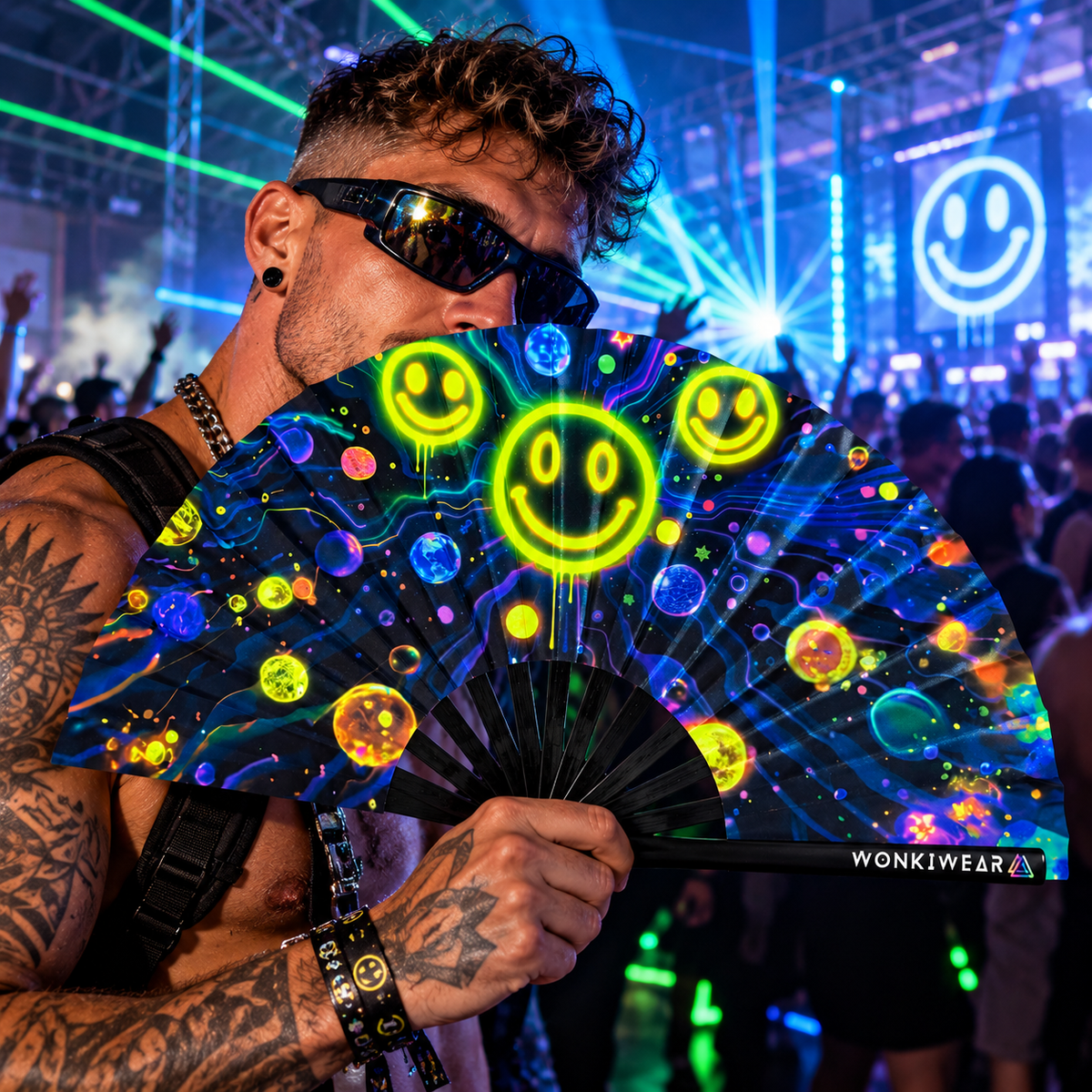 Male techno raver holding a WonkiWear galaxy smiley folding fan at a neon-lit UK rave.